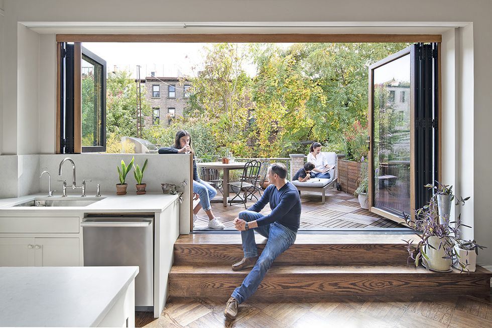 a family hangs out on the back terrace of their house, separated from the kitchen by a folding glass door