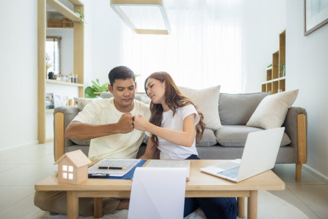 A man and a woman join fists while sitting in front of a coffee table with a computer, forms, a clipboard, and a small model house on it.