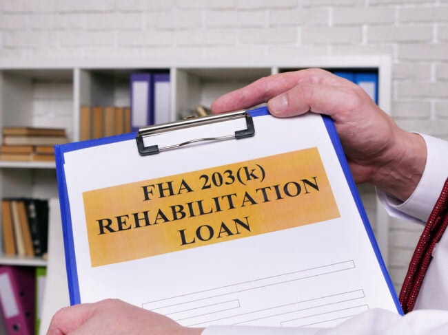 Two hands hold up a form on a clipboard with the words 'FHA 203(k) rehabilitation loan' on it.