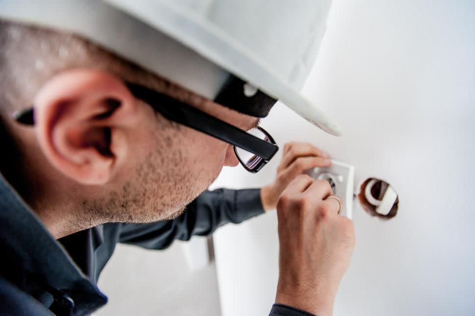 A man wearing a hard hat is diligently fixing a light switch on a white wall. His focused expression shows his concentration on the task at hand.